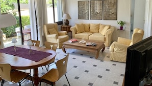 Un salon avec un canapé beige, une table basse en bois et un téléviseur. On y trouve deux lampes, une table avec des chaises et une plante en pot.