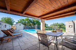 An outdoor pool area with lounge chairs and a table, surrounded by stone walls and trees.