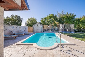 A rectangular swimming pool with a stone border, surrounded by a stone patio. There are lounge chairs and a small wooden structure in the background.