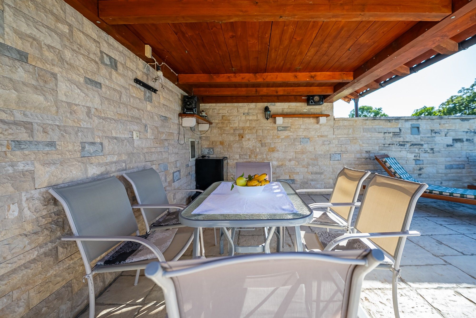 An outdoor patio with a stone wall, a wooden ceiling, and a table with chairs.