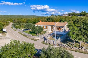 A stone house with a tiled roof, surrounded by greenery and a winding road.