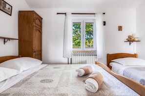 A room with two wooden beds, a large window with curtains, a wooden wardrobe, and a small shelf with a lamp and a teddy bear.