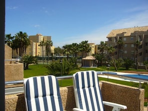 Un balcon avec deux fauteuils de jardin à rayures, une piscine et une vue sur un quartier résidentiel, avec des palmiers et des immeubles d’appartements.
