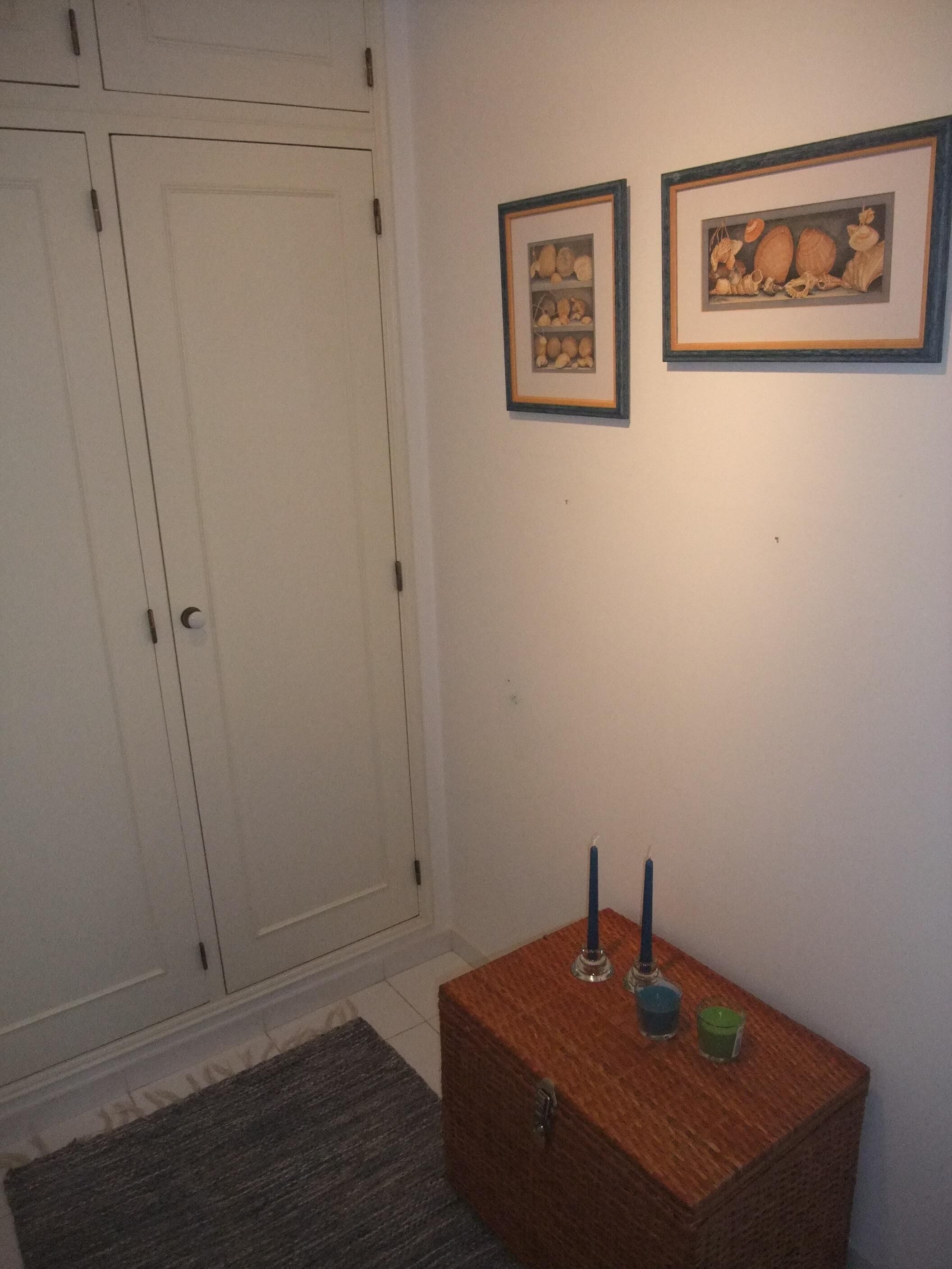 Una habitación con un armario blanco, un pequeño aparador de madera con velas, y dos cuadros en la pared.