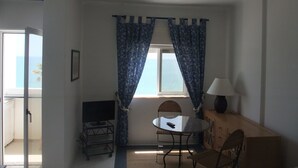 Ein Zimmer mit Fernseher auf einem Schrank, einem Tisch mit Stühlen, einer Lampe und Meerblick durch Vorhänge.