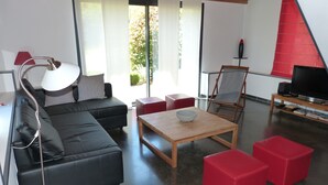 Una sala de estar moderna con un sofá de cuero negro, una mesa de centro de madera, otomanas rojas, una lámpara de pie y un televisor sobre un mueble de madera.