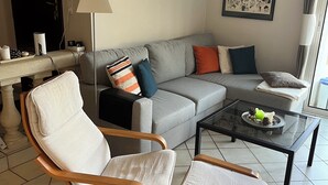 Een woonkamer met een grijze bank, twee witte fauteuils, een glazen salontafel en een ingelijste foto aan de muur.