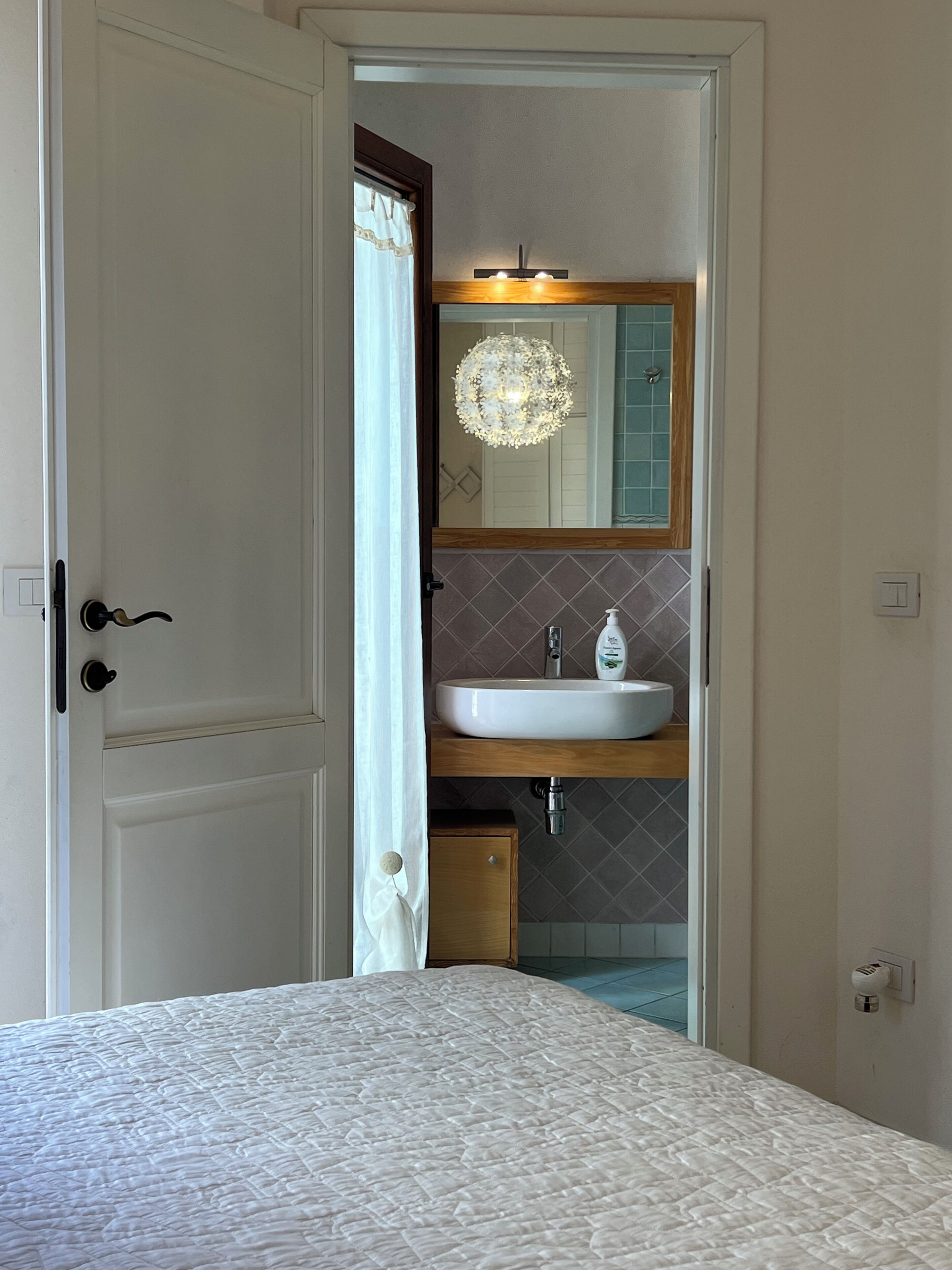 A bedroom with a bed, a bathroom with a sink and mirror, and a door leading to another room.