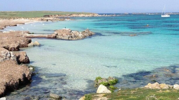 Een kustlandschap met helder turquoise water, rotsachtige rotsformaties en een verre zeilboot.