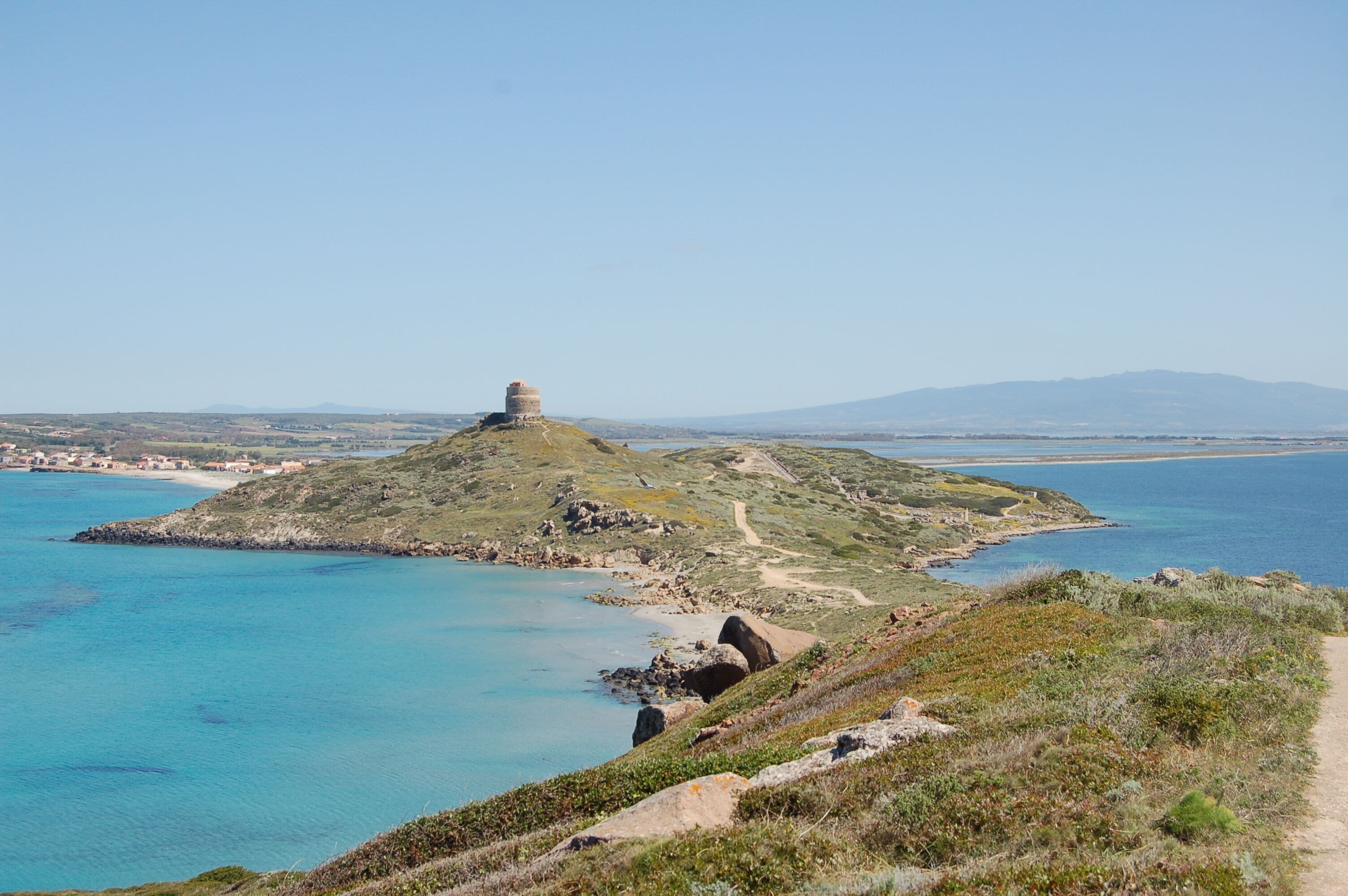 A coastal landscape with a prominent hill featuring a tower, clear blue waters, and a distant mountain range.