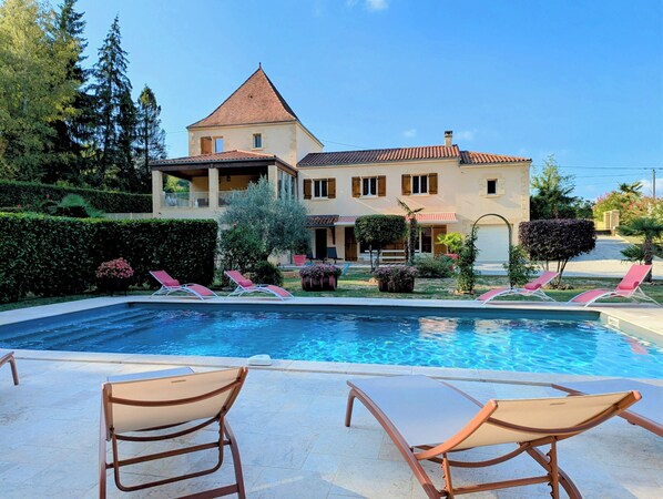 A swimming pool with lounge chairs, a large house with a balcony, and well-maintained gardens.