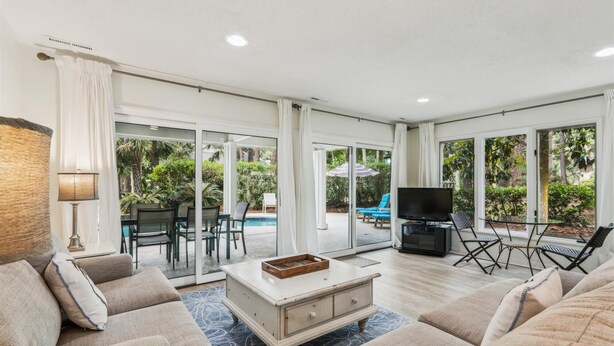 Un salon spacieux comprenant un ensemble de canapés, une table basse, un téléviseur et une cheminée. Une porte vitrée donne accès à un patio avec une piscine et du mobilier d’extérieur.