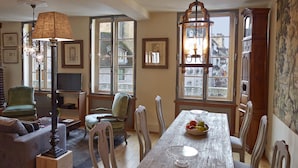 Uma sala de jantar com uma mesa de madeira, cadeiras brancas e um candelabro. Há uma poltrona verde e um sofá ao fundo.