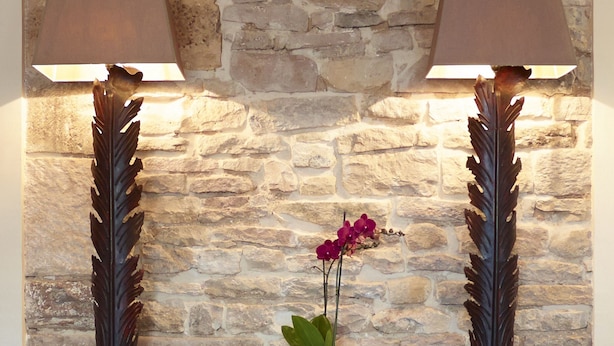 Uma parede de pedra com uma mesa de consola de madeira, duas lâmpadas, uma planta em vaso e uma cadeira de vime.