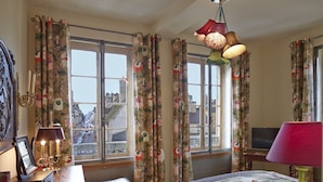 Quarto com cortinas florais, uma cama, uma mesa de escritório com abajur e vista da cidade através de grandes janelas.
