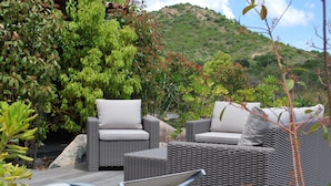 An outdoor seating area with wicker furniture and cushions, surrounded by lush greenery and a mountainous backdrop. - Walk-new in charming residence with 300m² swimming pool in Porto Vecchio (Porto-Vecchio)