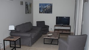 A living room with a grey sofa, a coffee table, a TV on a stand, and a lamp on a side table. - Walk-new in charming residence with 300m² swimming pool in Porto Vecchio (Porto-Vecchio)