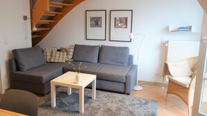 Een woonkamer met een grijze bank, een witte salontafel, een rieten fauteuil en een trap.