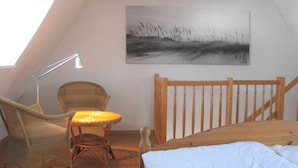 Een knus slaapkamer onder het dak, met een schuine wand, een rieten stoel, een klein tafeltje, een houten bed en een schilderij aan de muur.