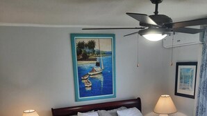 Un dormitorio con cama, dos lámparas de noche, ventilador de techo, un cuadro de barcos y una fotografía enmarcada en la pared.