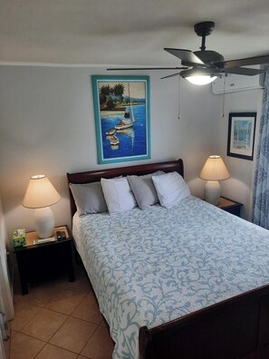 A bedroom with a bed, two bedside lamps, a ceiling fan, a painting of boats, and a framed picture on the wall.
