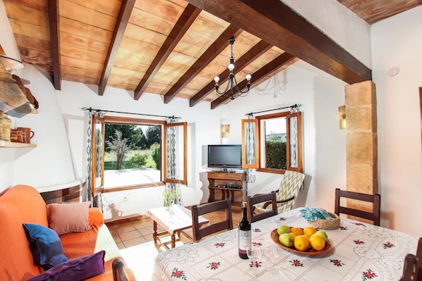 Um quarto acolhedor com teto de madeira, uma mesa de jantar com uma garrafa de vinho e frutas, e uma televisão ao fundo.