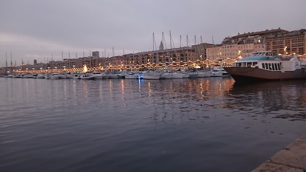 Un puerto deportivo con barcos amarrados, un paseo marítimo y edificios al fondo.