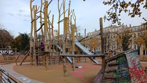 Un parque infantil con estructuras para escalar y toboganes, rodeado de una zona de arena y un camino pavimentado.