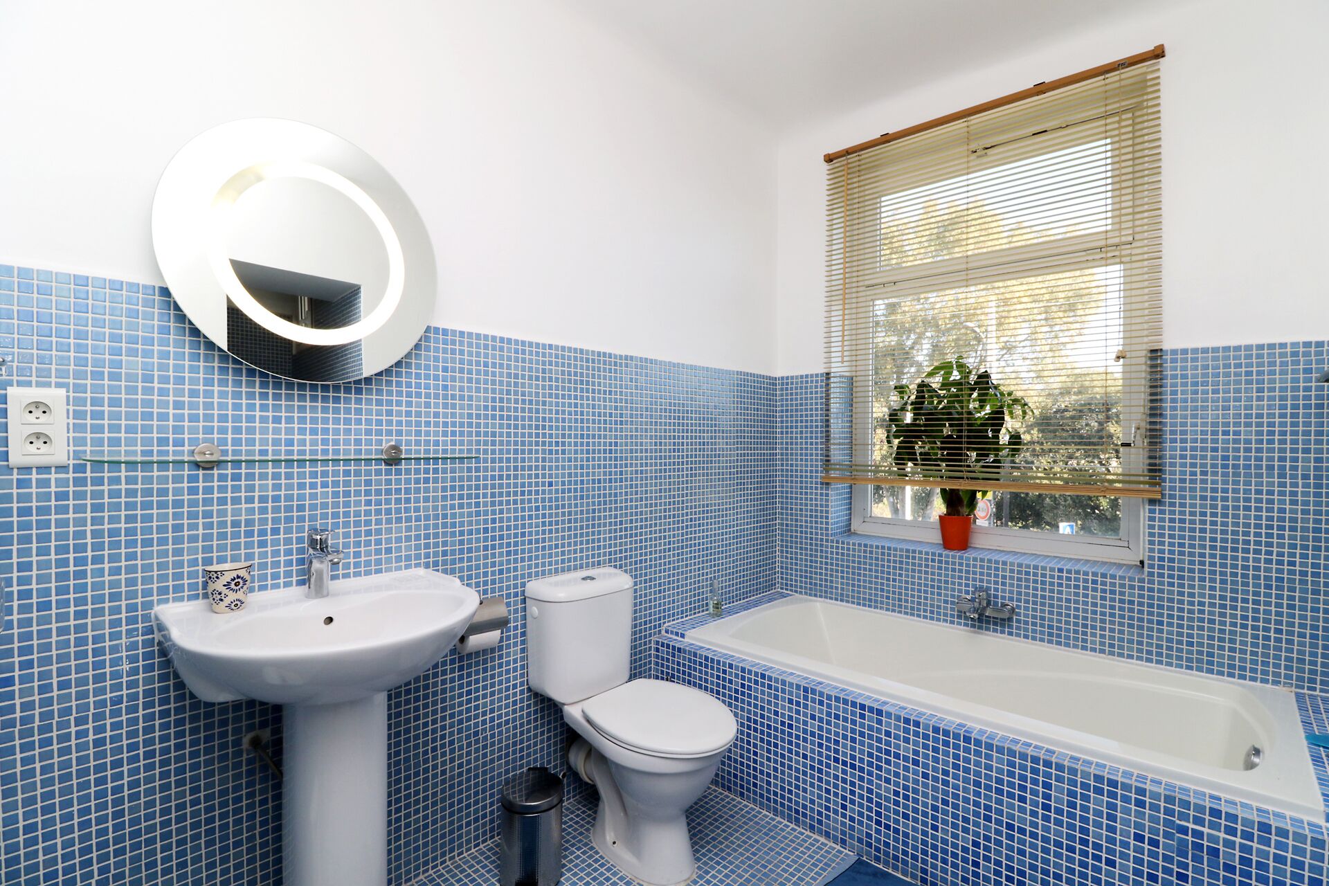 Un baño con paredes de azulejos azules, un lavamanos blanco, un inodoro, una bañera, un espejo y una ventana con persianas.