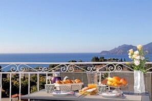 Una mesa en el balcón con alimentos para el desayuno, un jarrón de flores y una vista panorámica del mar y las montañas.