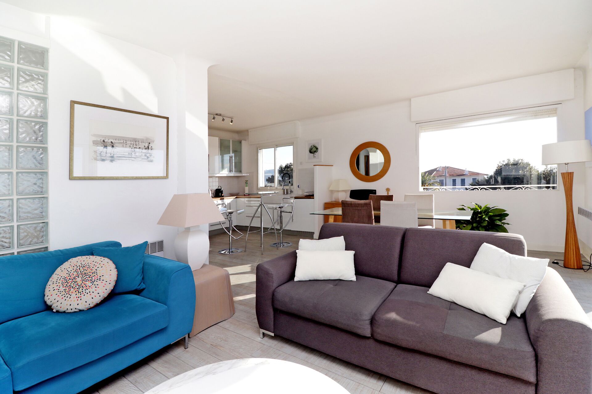 A modern living room with a blue sofa and a grey sofa, a dining area with a table and chairs, a kitchen in the background, and a large window with a view.
