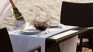 Mesa lista para dos con una botella de champán, una jarra de vidrio y un florero con flores en un entorno playero.