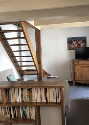 Een trap met een houten leuning die uitkomt op een plateau. Op de voorgrond staat een boekenplank vol boeken. Een televisie staat op een houten kast aan de rechterkant.