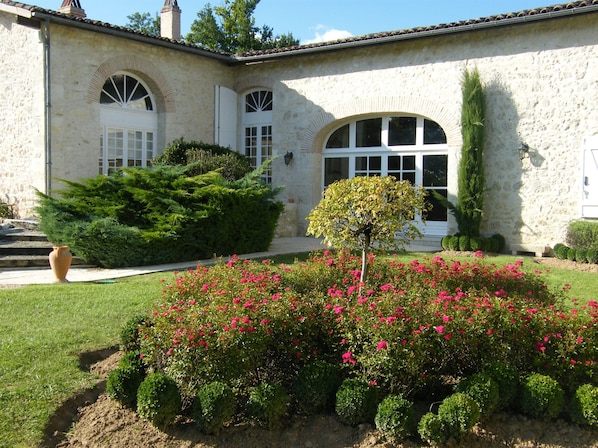 Un bâtiment à la façade en pierre, doté de fenêtres en arc de cercle et agrémenté d’un jardin bien entretenu, où s’épanouissent des fleurs roses et où les arbustes sont taillés avec soin.