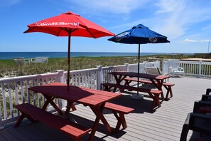 Une terrasse en bois avec des tables de pique-nique et des bancs, des parasols rouges et bleus, et une vue sur l’océan.