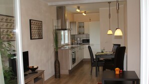 A modern kitchen with wooden cabinets, a stainless steel range hood, and pendant lighting. There is a dining area with a table and chairs, and a television on a stand.