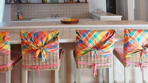 A dining area with a thatched ceiling, wicker chairs with colorful fabric, and a kitchen in the background.