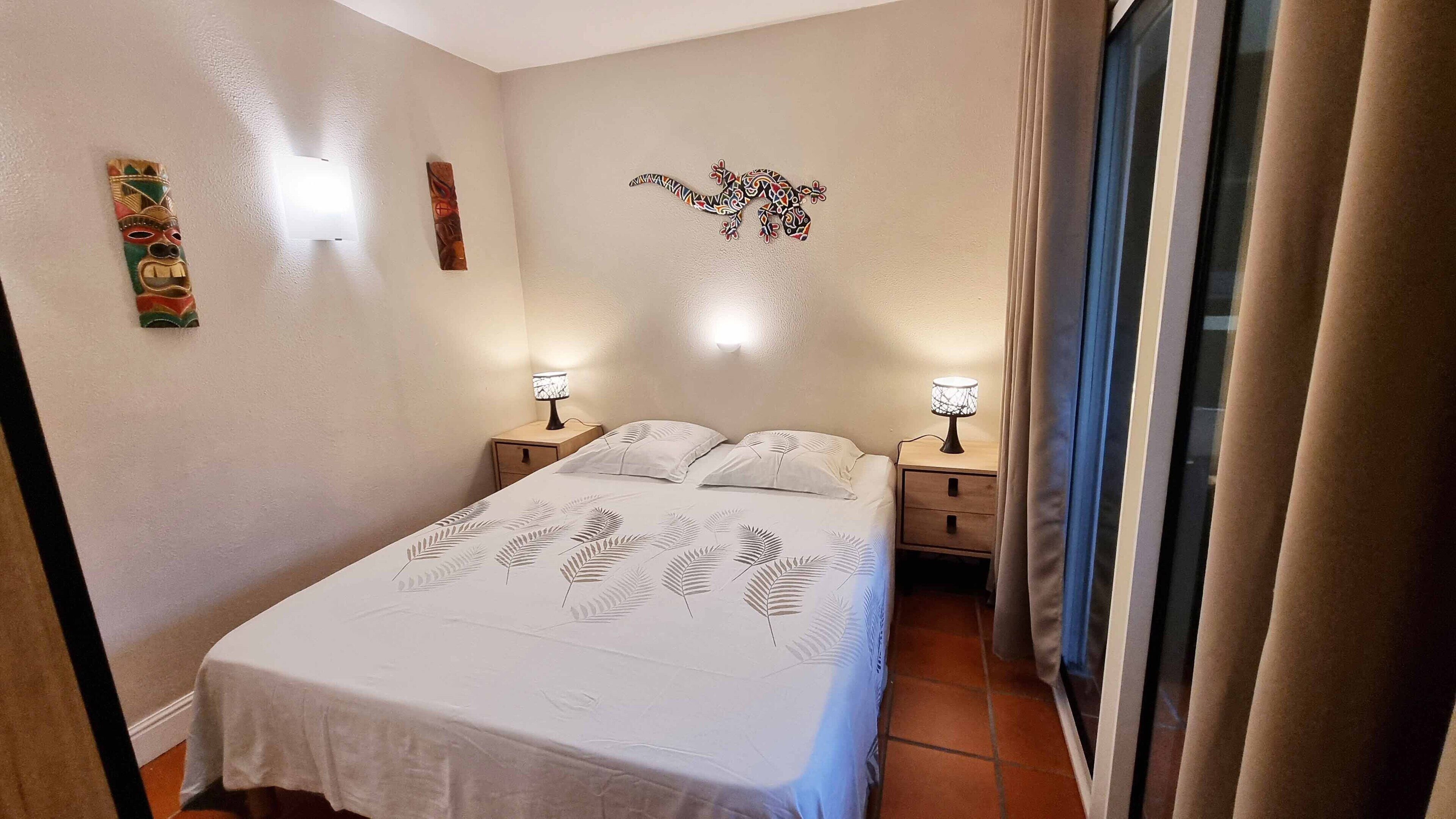 A bedroom with a bed, two bedside tables, a wall-mounted lamp, and a window with curtains.