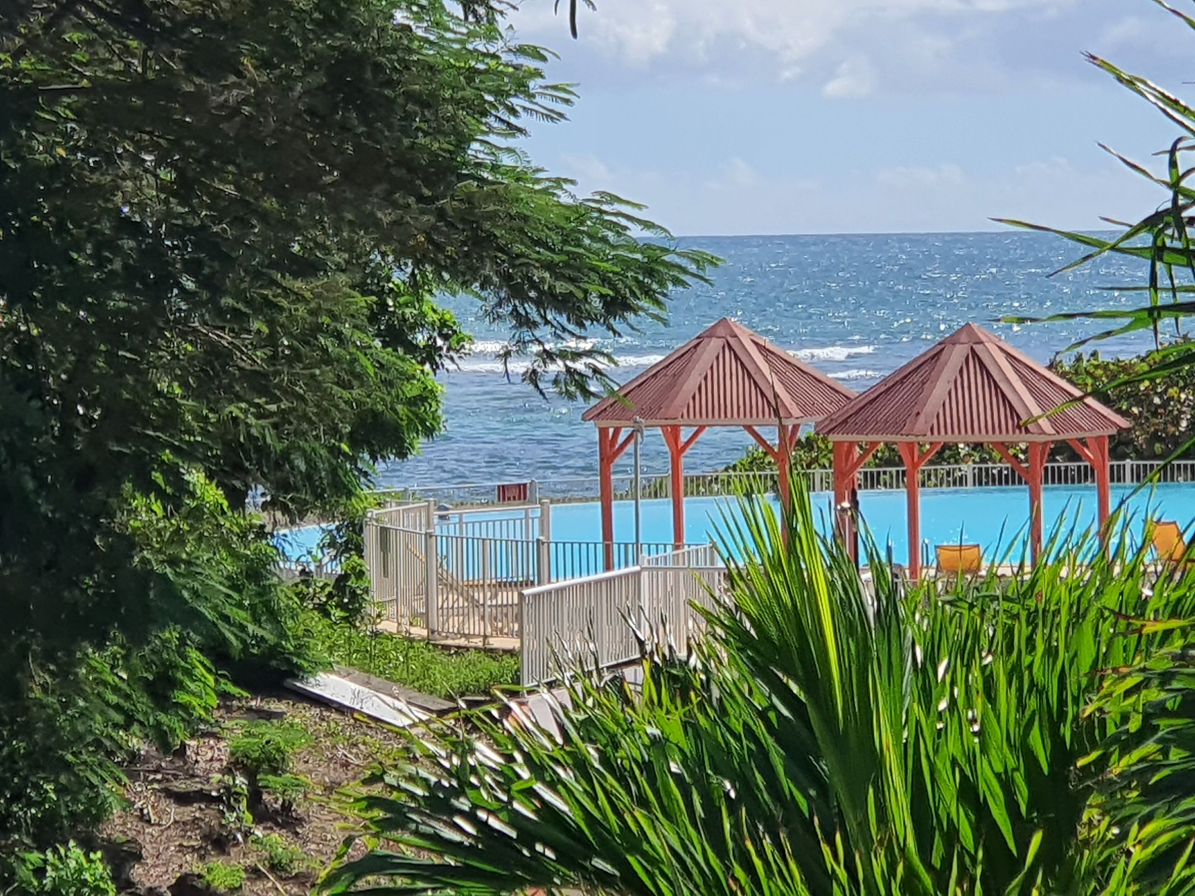 Vista desde la playa con dos gazebos de madera, una piscina y una vista despejada del océano.