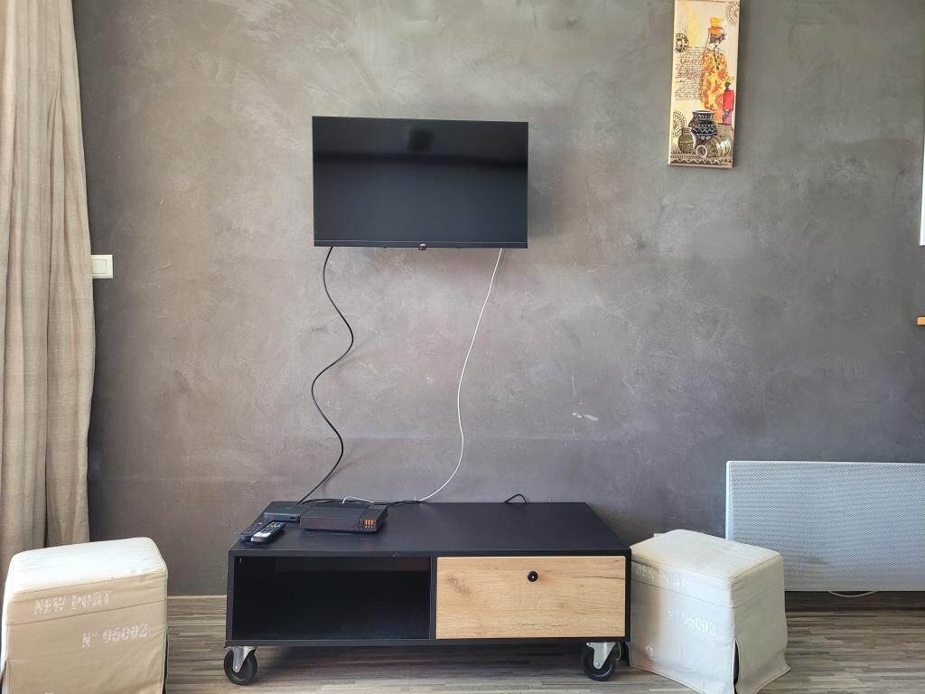 Habitació amb televisió penjada, un moble negre per a entreteniment amb un calaix, un otomà blanc i una obra d'art penjada a la paret.