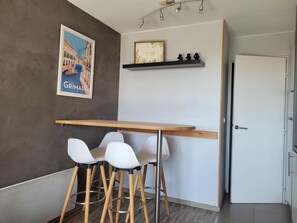 Une pièce avec une table en bois et quatre chaises blanches, un tableau encadré au mur, une étagère avec des objets décoratifs et une porte blanche.