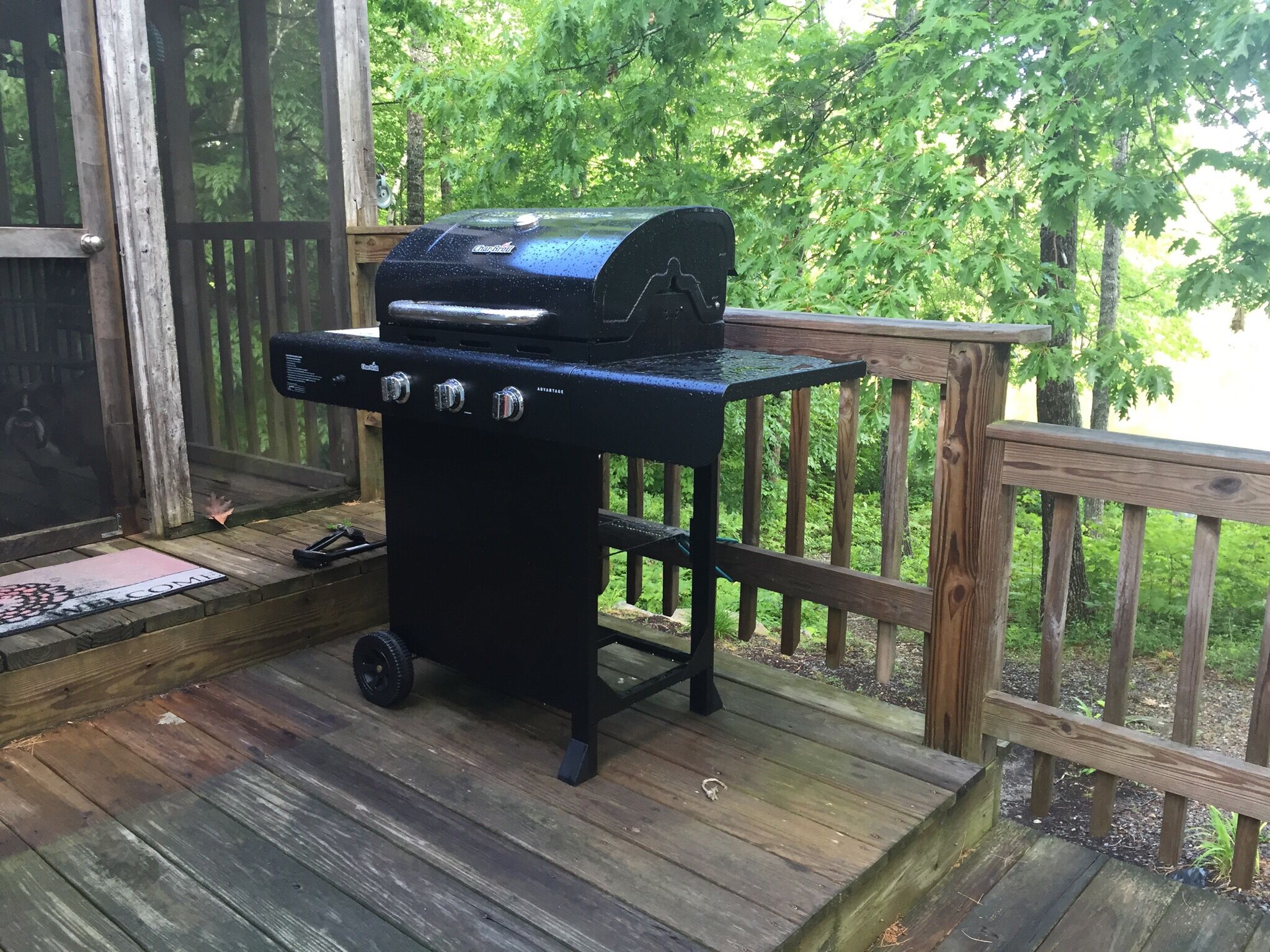 A black barbecue grill on a wooden deck with a wooden railing and greenery in the background.