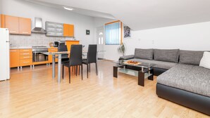 A modern kitchen with orange cabinets, a dining area with a glass table, and a living area with a gray sofa. - Apartment Tena (Ližnjan)