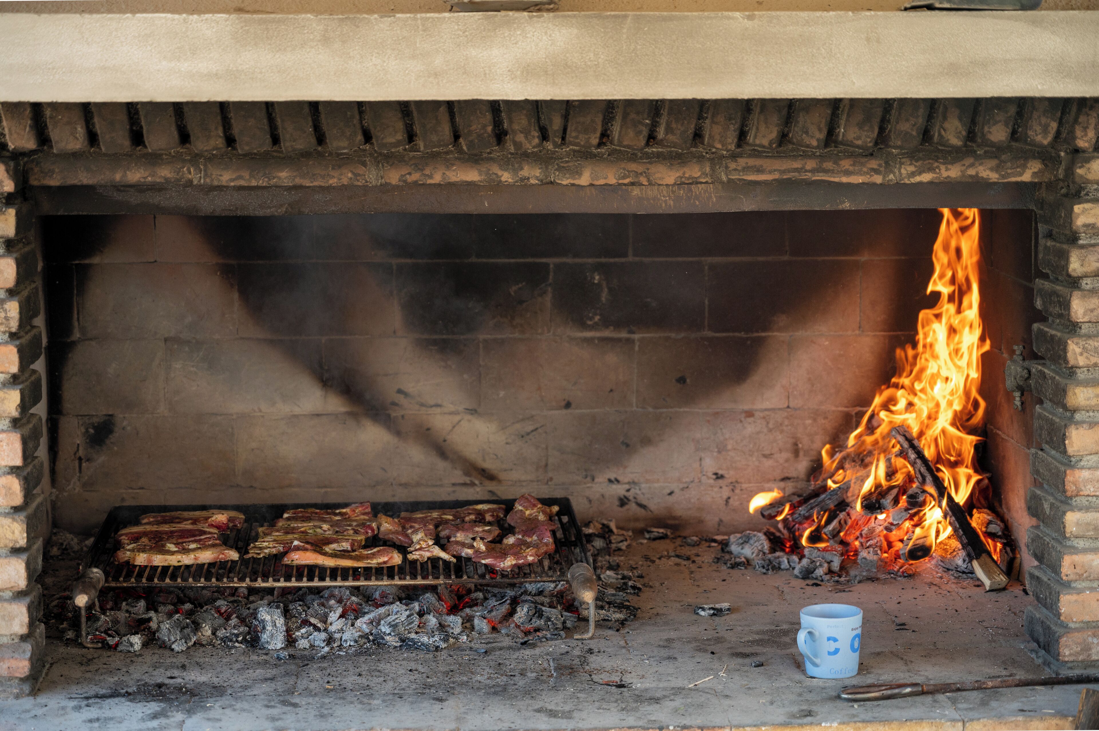 A brick fireplace with a fire burning, a grill with meat, and a mug on the floor.