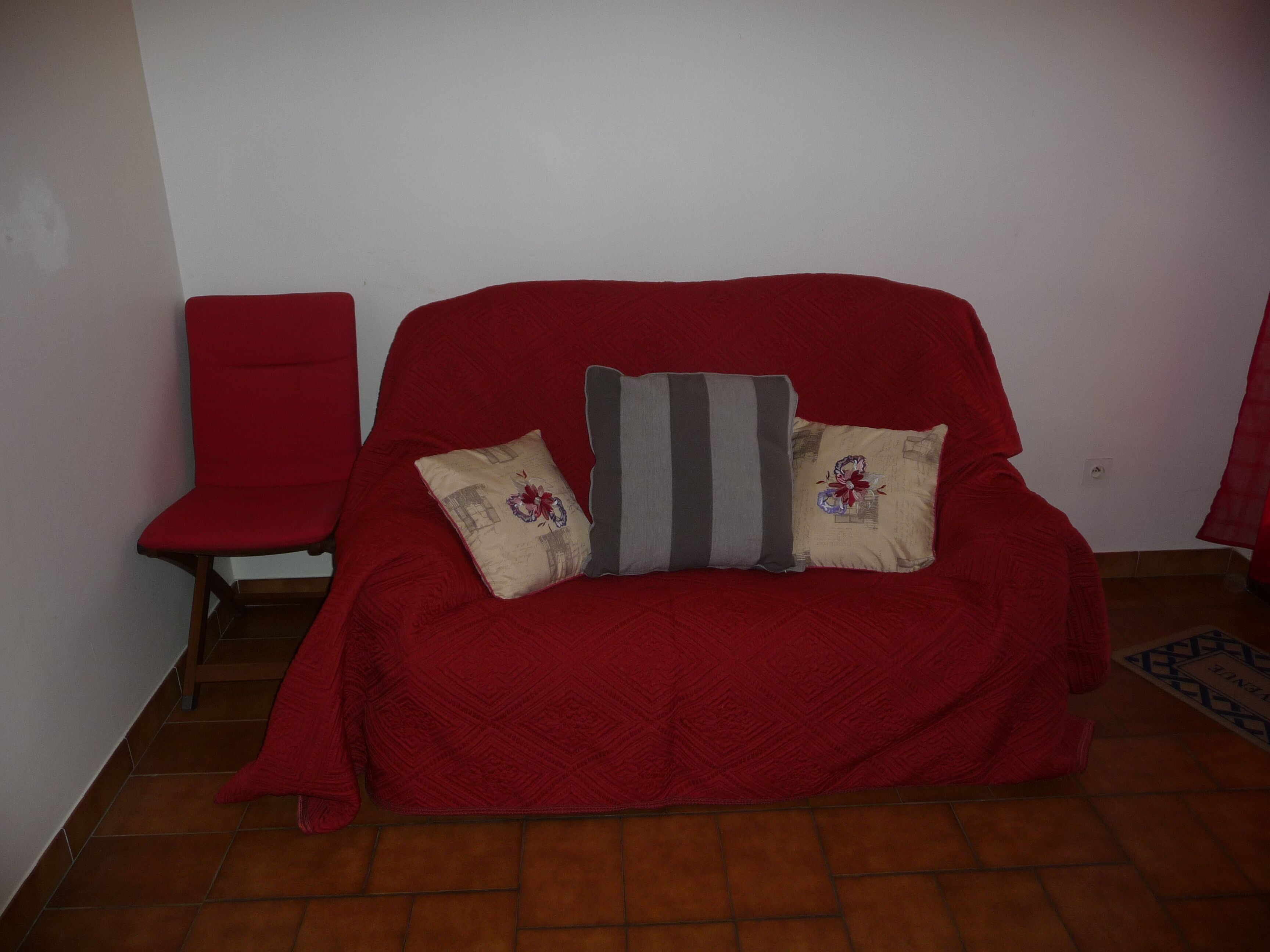 Um sofá vermelho com duas almofadas, uma cadeira vermelha e um tapete estampado sobre um piso de azulejos.