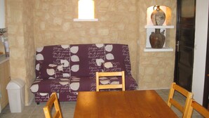 A cozy living room with a stone wall, a purple sofa with white patterns, a wooden dining table, and a small shelf with decorative items. - F3 feet in the water. 50 m from the beach (Sagone)
