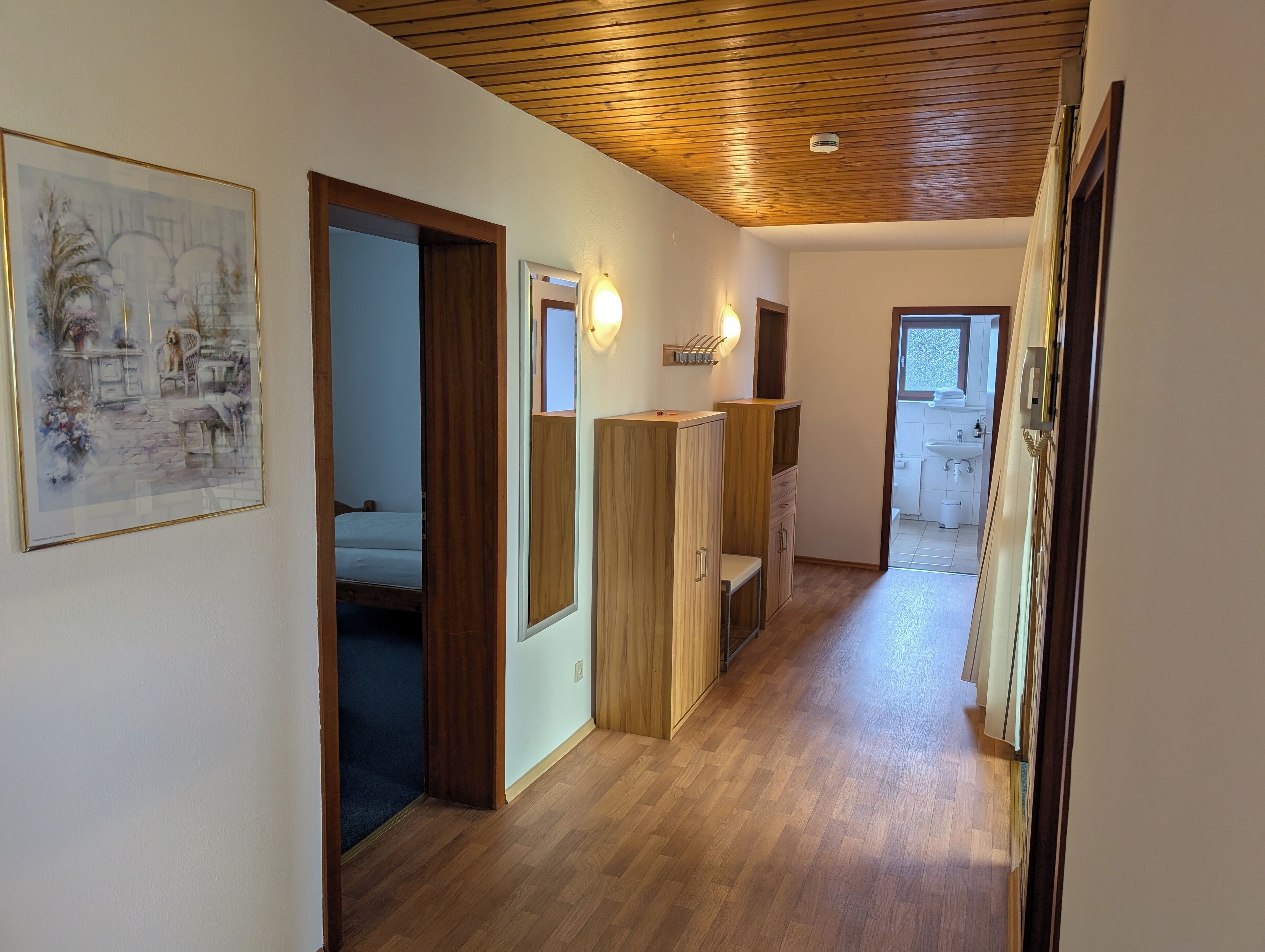 A hallway with wooden flooring, a bed in the adjacent room, a wooden wardrobe, and a bathroom visible through an open door.