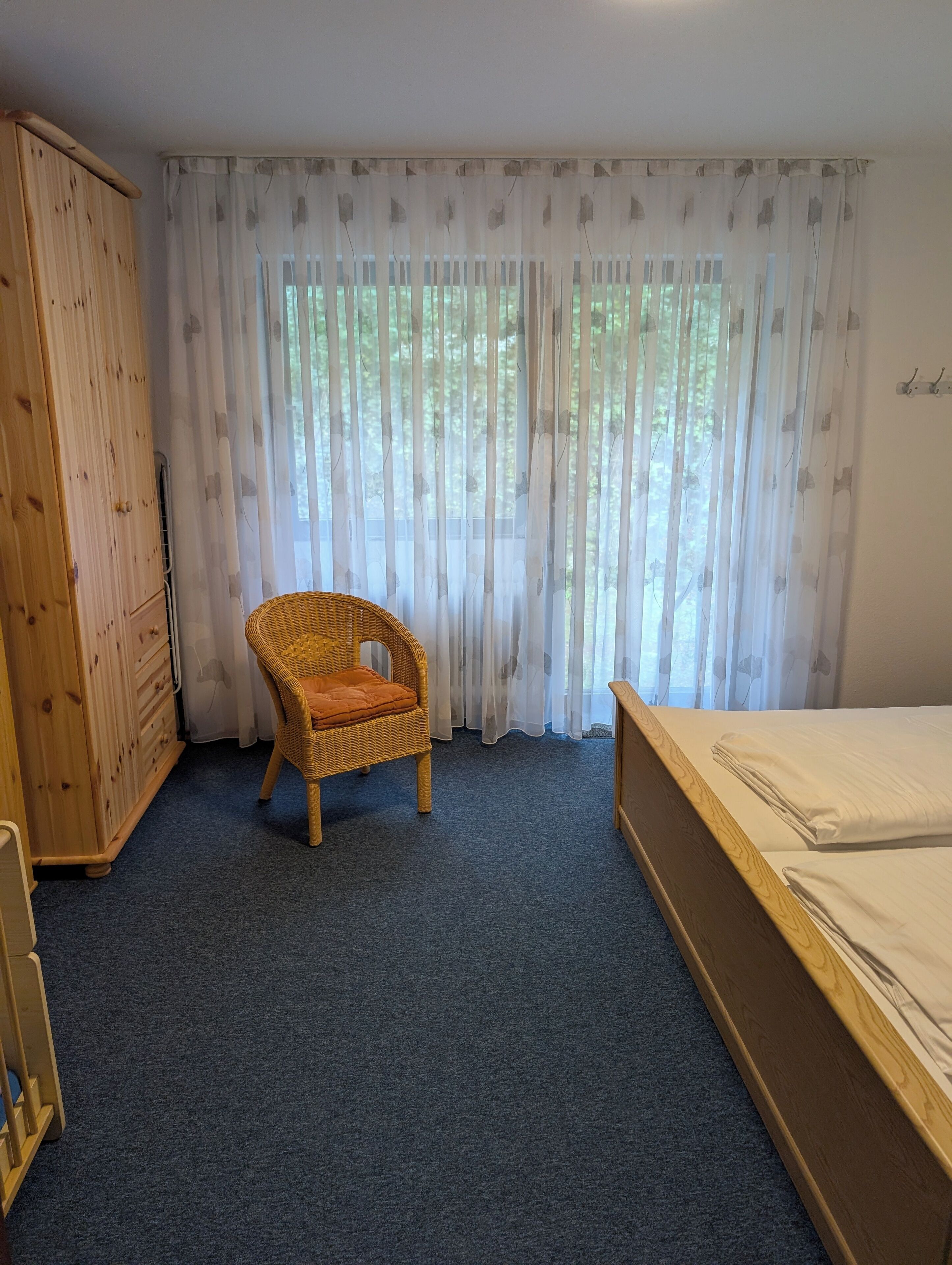 Une chambre avec une armoire en bois, une chaise en osier avec un coussin, un lit avec une literie blanche et des rideaux légers ornés d’un motif d’arbre.