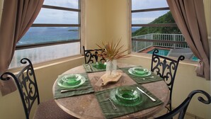 A dining table set with green glassware, a vase with dried plants, and a wooden serving board, overlooking a scenic view of the ocean and a lush landscape. - Casa de Suenos | St. John (Coral Bay)