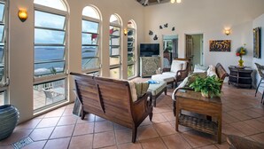 A spacious living room with large arched windows, a wooden bench, a coffee table, and a television. - Casa de Suenos | St. John (Coral Bay)